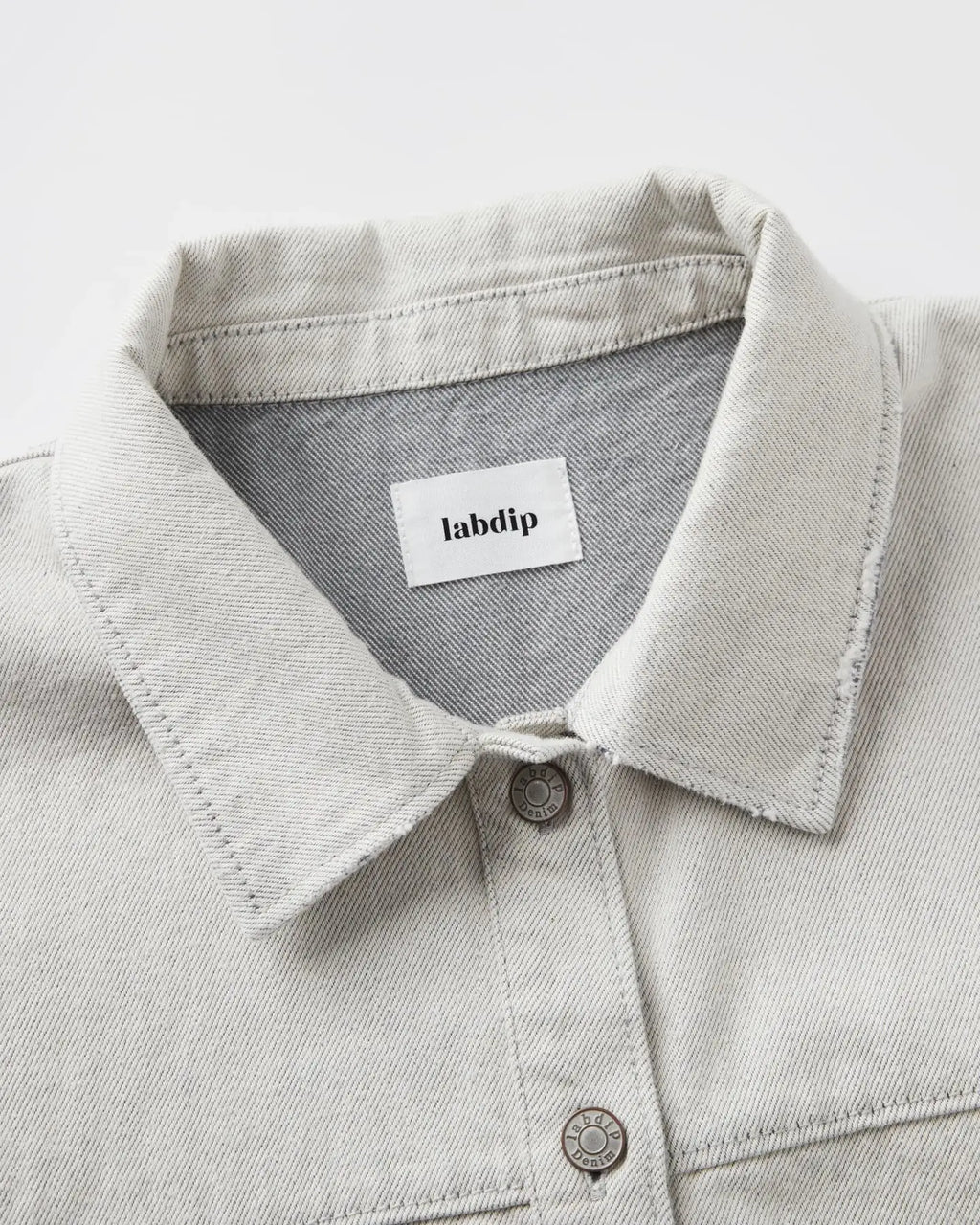 Col chemise en denim non teint couleur granit clair avec coutures apparentes et boutons métalliques, tissu 100% coton au toucher structuré et naturel.