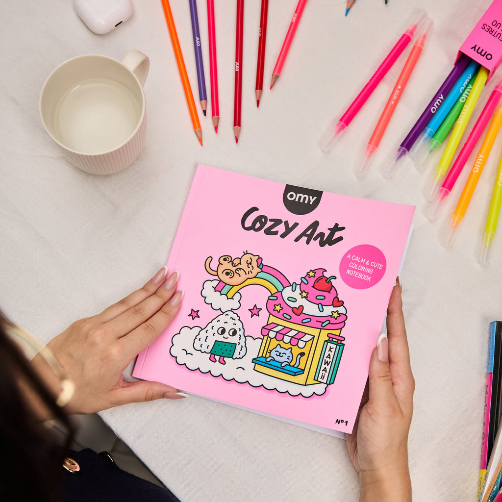 Main tenant le cahier de coloriage Cozy Art Kawaii d’OMY sur une table recouverte de feutres colorés, ambiance douce et créative.
