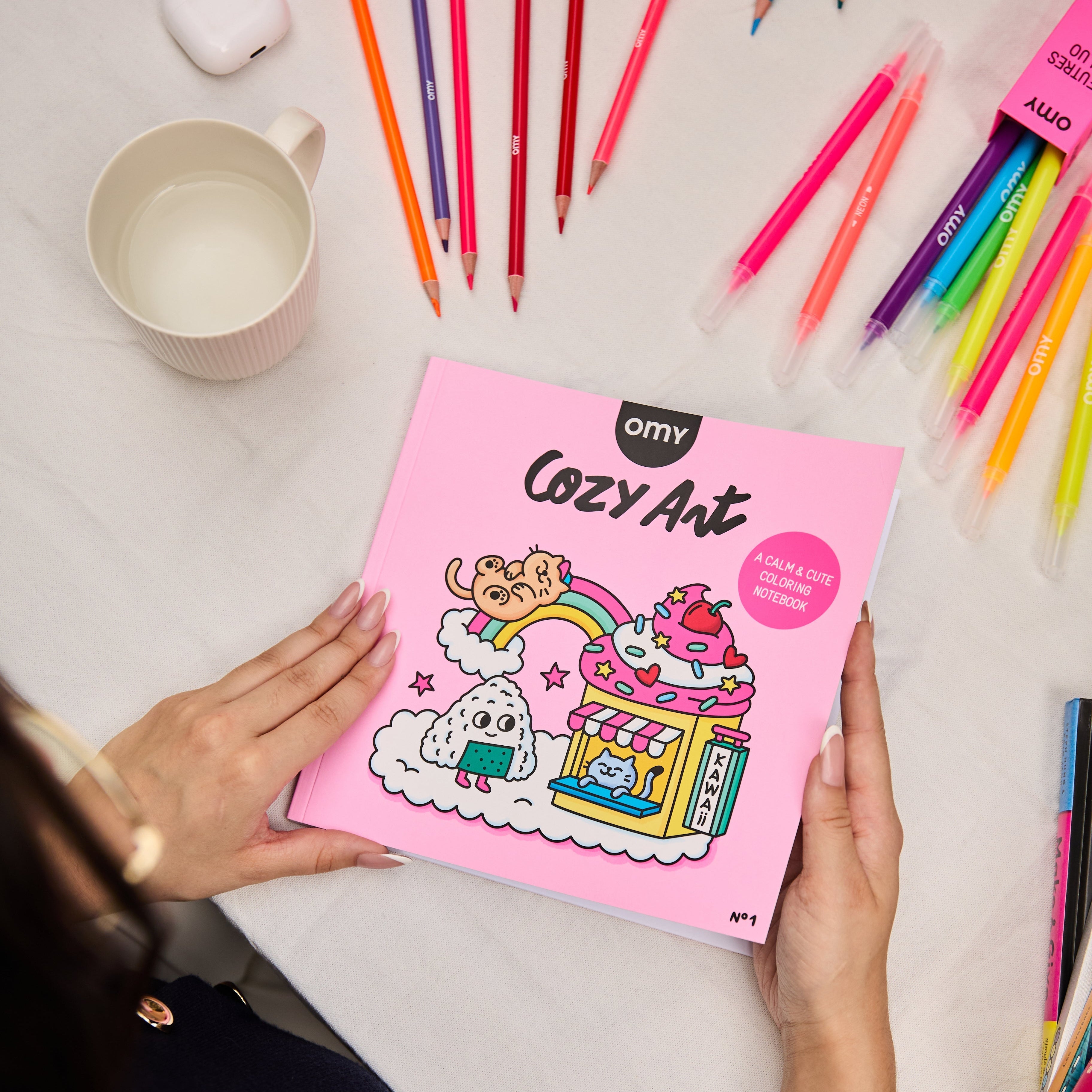 Main tenant le cahier de coloriage Cozy Art Kawaii d’OMY sur une table recouverte de feutres colorés, ambiance douce et créative.