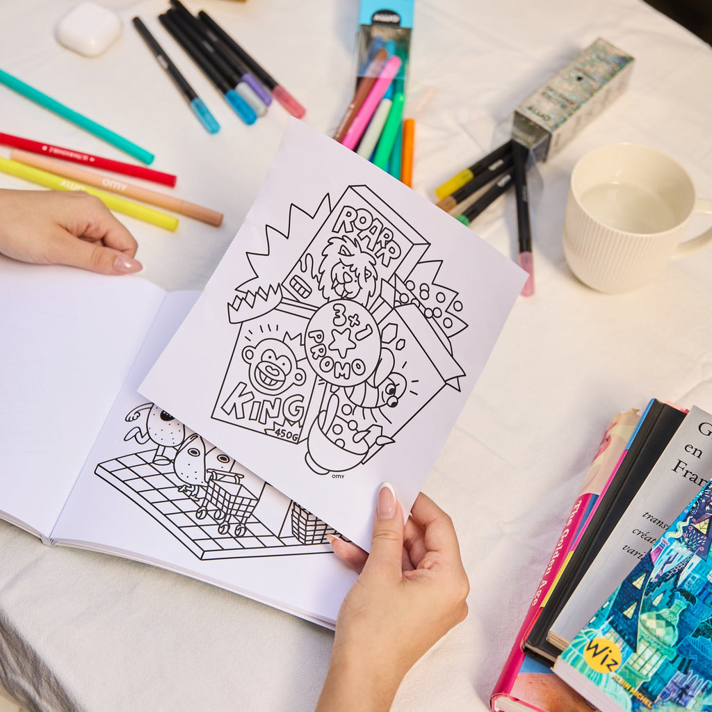 Page extraite du carnet Cozy Art Super Market avec illustration à colorier de produits alimentaires, tenue par une main au-dessus du carnet ouvert sur une table garnie de feutres et livres.
