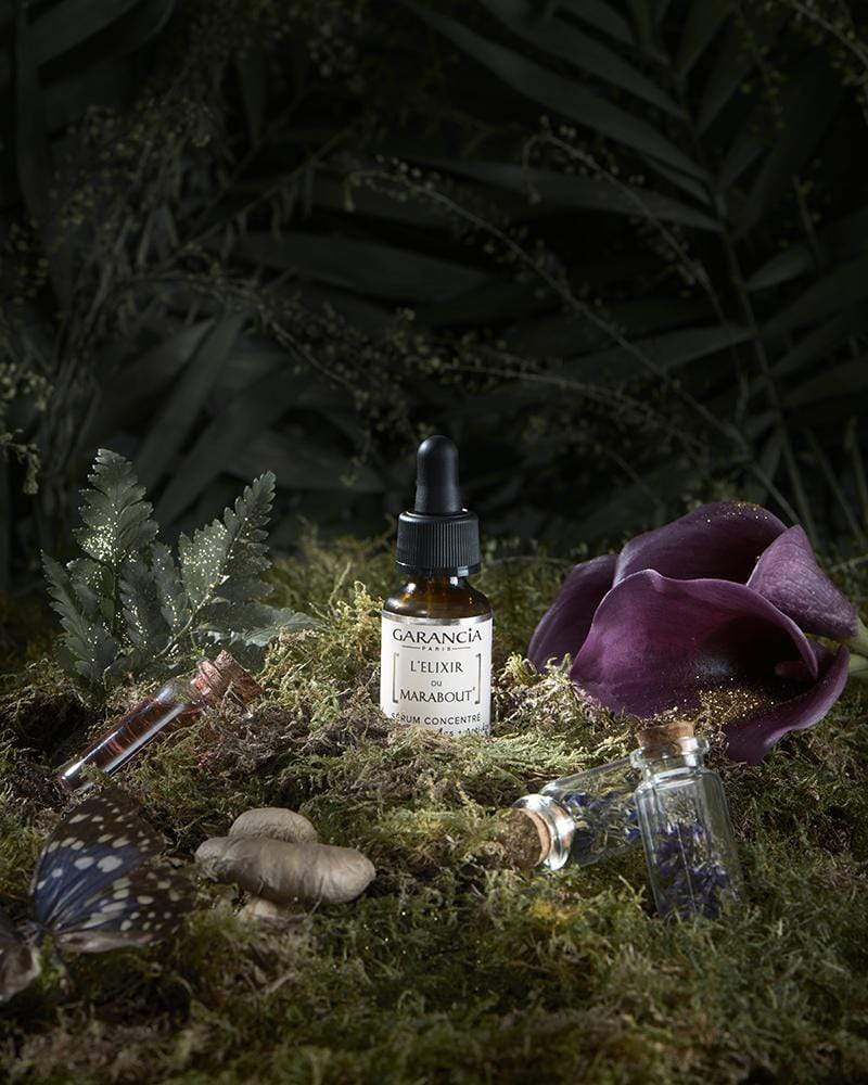 Sérum L'Élixir du Marabout de Garancia présenté dans un décor de forêt enchantée avec de la mousse, des fleurs violettes, un papillon et des fioles magiques.