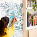 Mise en ambiance du rouleau tropical OMY collé sur un mur blanc, entre bibliothèque et plante verte
