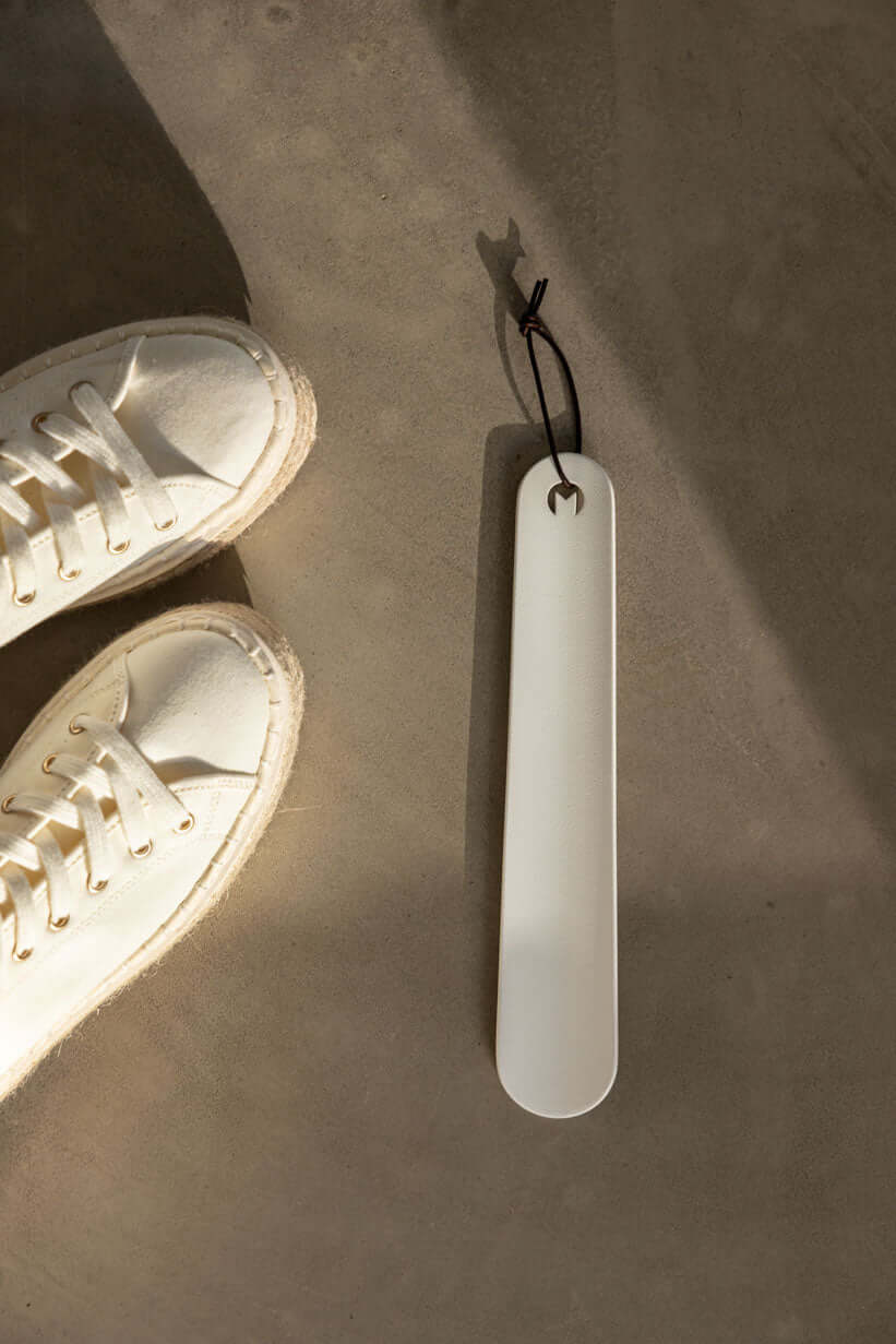 Schuhlöffel TILO aus Metall neben weißen Sneakern auf einem modernen Untergrund.