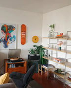 Stylish living room setup featuring a vibrant Warhol Campbell’s Soup Peach skateboard deck mounted on the wall, accompanied by modern art prints, lush green plants, and minimalist shelving adorned with books, decor items, and a turntable for a cozy and creative atmosphere.