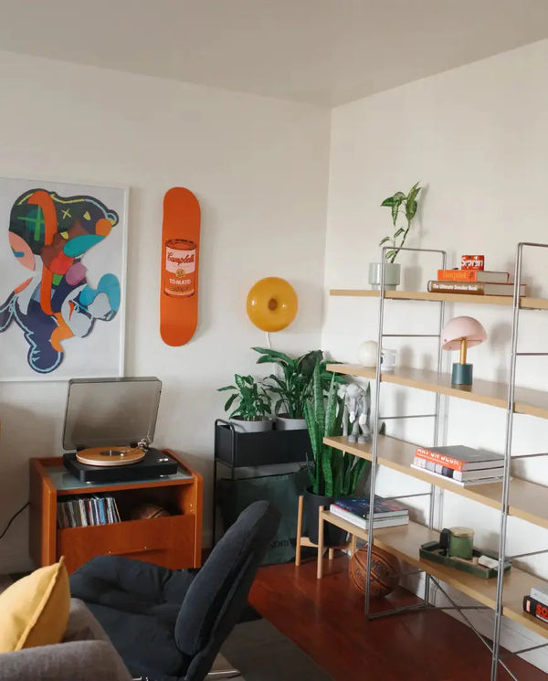 Stylish living room setup featuring a vibrant Warhol Campbell’s Soup Peach skateboard deck mounted on the wall, accompanied by modern art prints, lush green plants, and minimalist shelving adorned with books, decor items, and a turntable for a cozy and creative atmosphere.