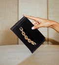 Black silk satin Catena Envelope Clutch with gold chain embroidery held against a textured backdrop.