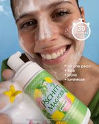 Une femme souriante applique la mousse onctueuse du Pschitt Magique Garancia sur son visage. Elle tient le flacon édition limitée devant un ciel bleu parsemé de nuages, illustrant un moment de soin frais et naturel.